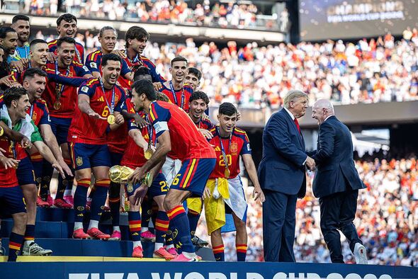 A soccer team is celebrating in the foreground on the field, with a large crowd of spectators in the background. Two individuals