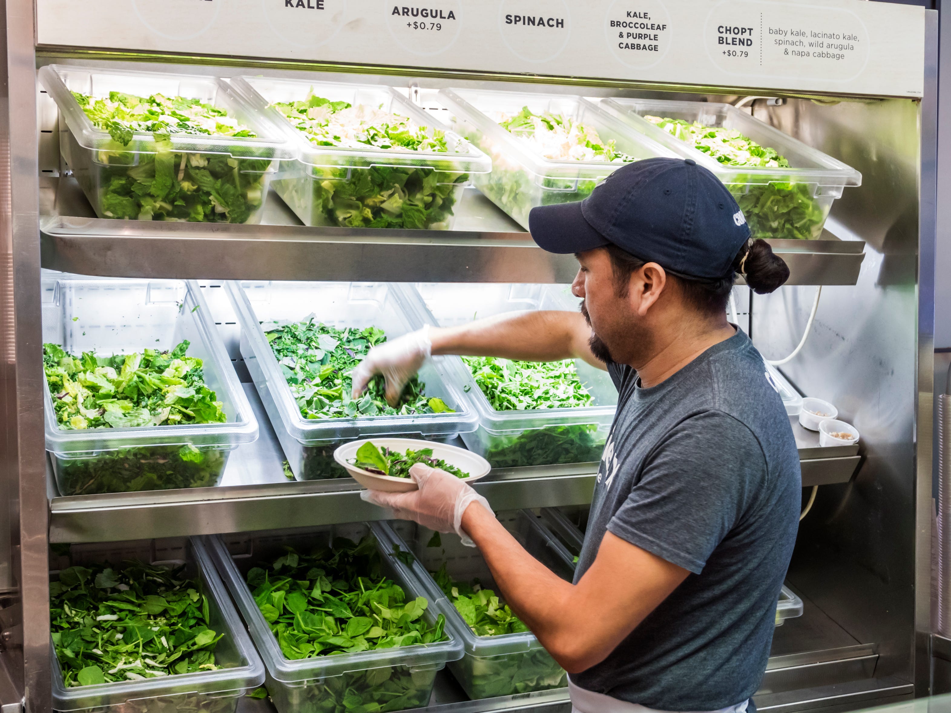 An employee at a Chopt restaurant picks leafy greens from a plastic container and places them into a bowl behind the counter while wearing a baseball hat.