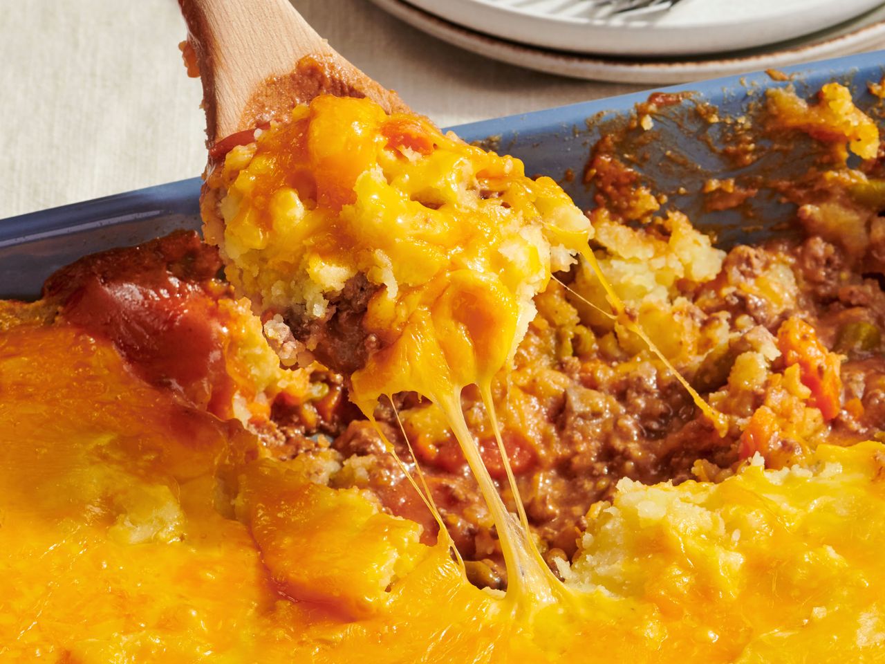 A casserole dish with a serving of cheesy layered casserole being lifted with a wooden spoon