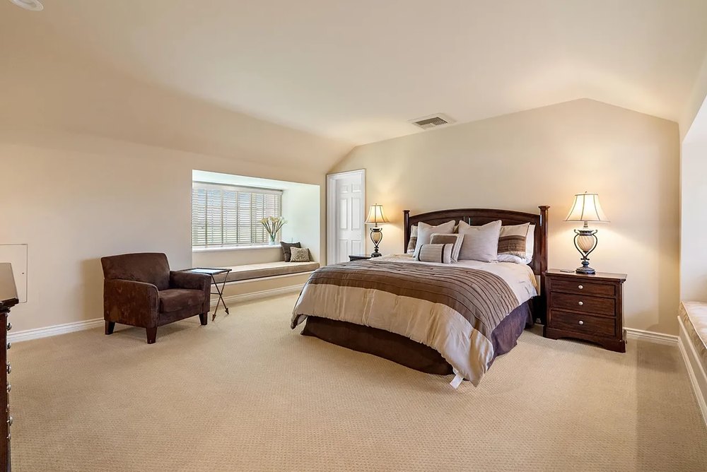 Primary bedroom suite with neutral carpeting, dark wood nightstands, and a built-in window seat.