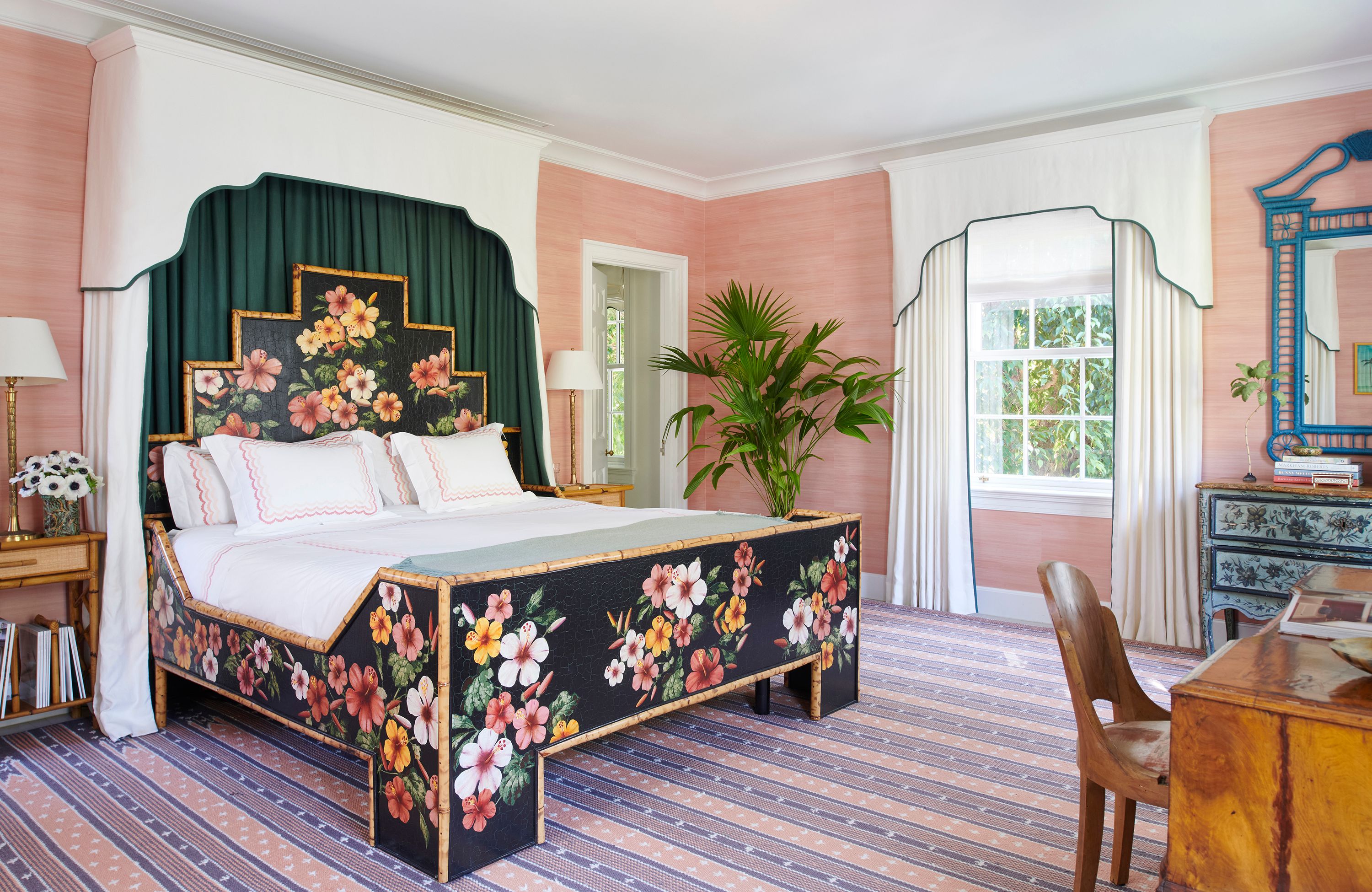 One guest bedroom boasts a lively, painted, bamboo bed frame ensconced in white curtains that match the window dressings. The nightstands also incorporate bamboo. Striated pink walls pick up the stripes in the pink and purple rug, which complements the blue of a nearby rattan mirror.