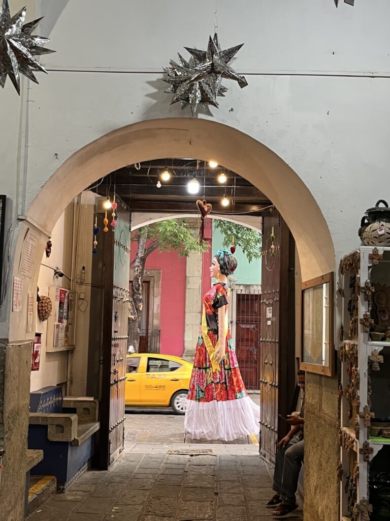 A colorful traditional Oaxacan muñeca wearing a vibrant embroidered dress hangs beneath an arched doorway inside a shop. Through the open entrance, a yellow taxi and colorful colonial buildings are visible across the street.