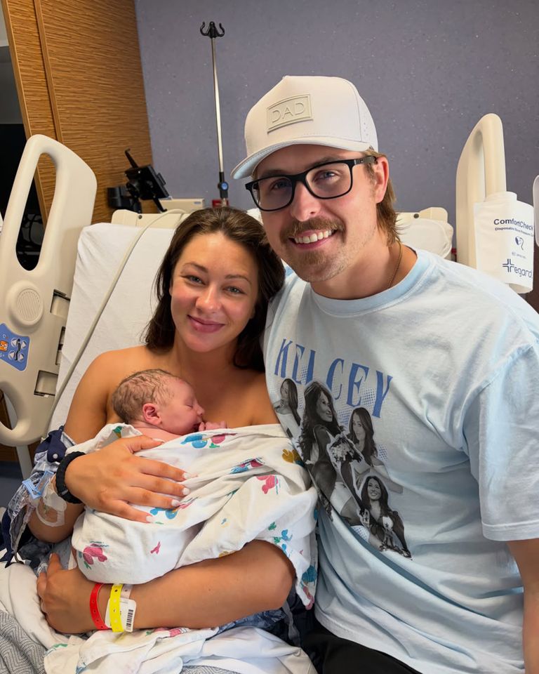 A woman holding a newborn baby next to a man wearing glasses and a cap in a hospital room