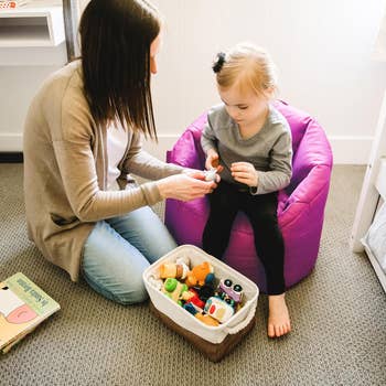 Woman and young child sitting together on carpet; child sits on bean bag, exploring toys from a storage basket. Educational play setting