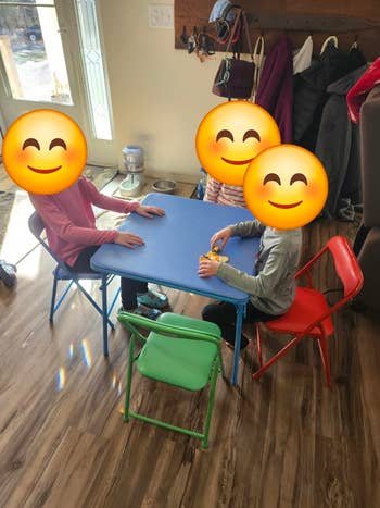 Three children sit at a small table in a cozy room, playing with toys. Bright chairs contrast with the wooden floor, creating an inviting play area