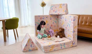 Child plays in a cushioned play structure with patterned fabric, surrounded by stuffed animals and toys, in a cozy room setting