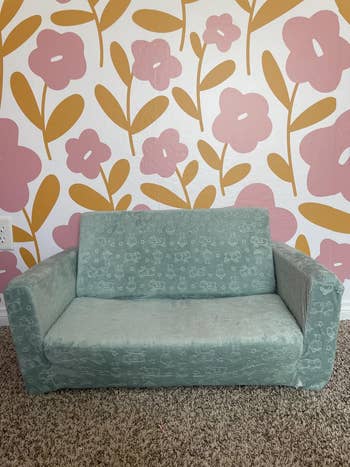 Child-sized sofa with a pattern of small, playful drawings, set against a floral patterned wall. Ideal for a kid's reading nook