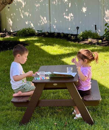 Two children sit at a small picnic table outdoors, engaged in painting with watercolor sets on a sunny day