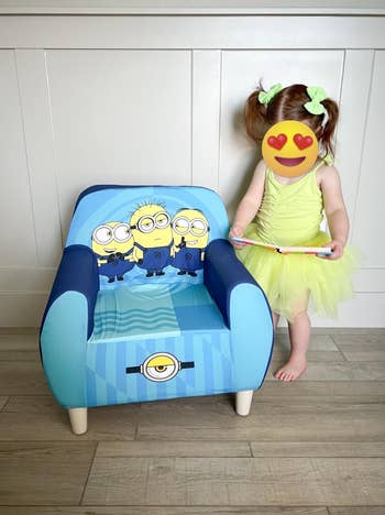 Child with a blurred face in a green dress stands beside a small chair featuring Minions graphics