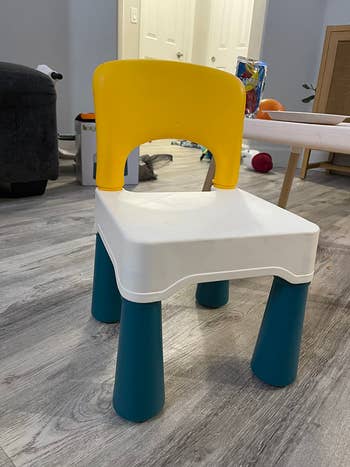 Children's chair with a yellow backrest, white seat, and teal legs, placed on a wood floor in a living room setting