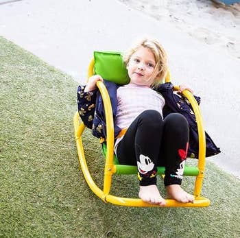 Child sitting in a yellow outdoor rocker chair on grass, wearing leggings with animated character design