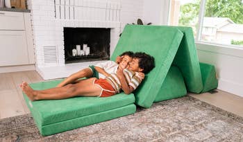 Two children smiling and lounging on a foldable green play couch in a cozy living room, near a fireplace