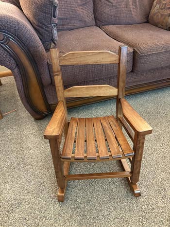 A small wooden rocking chair with slatted back and seat, placed on a carpeted floor in front of a sofa