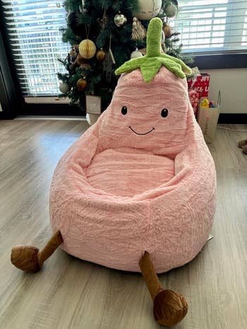 Plush armchair shaped like a smiling eggplant with a green top, displayed indoors near a decorated Christmas tree