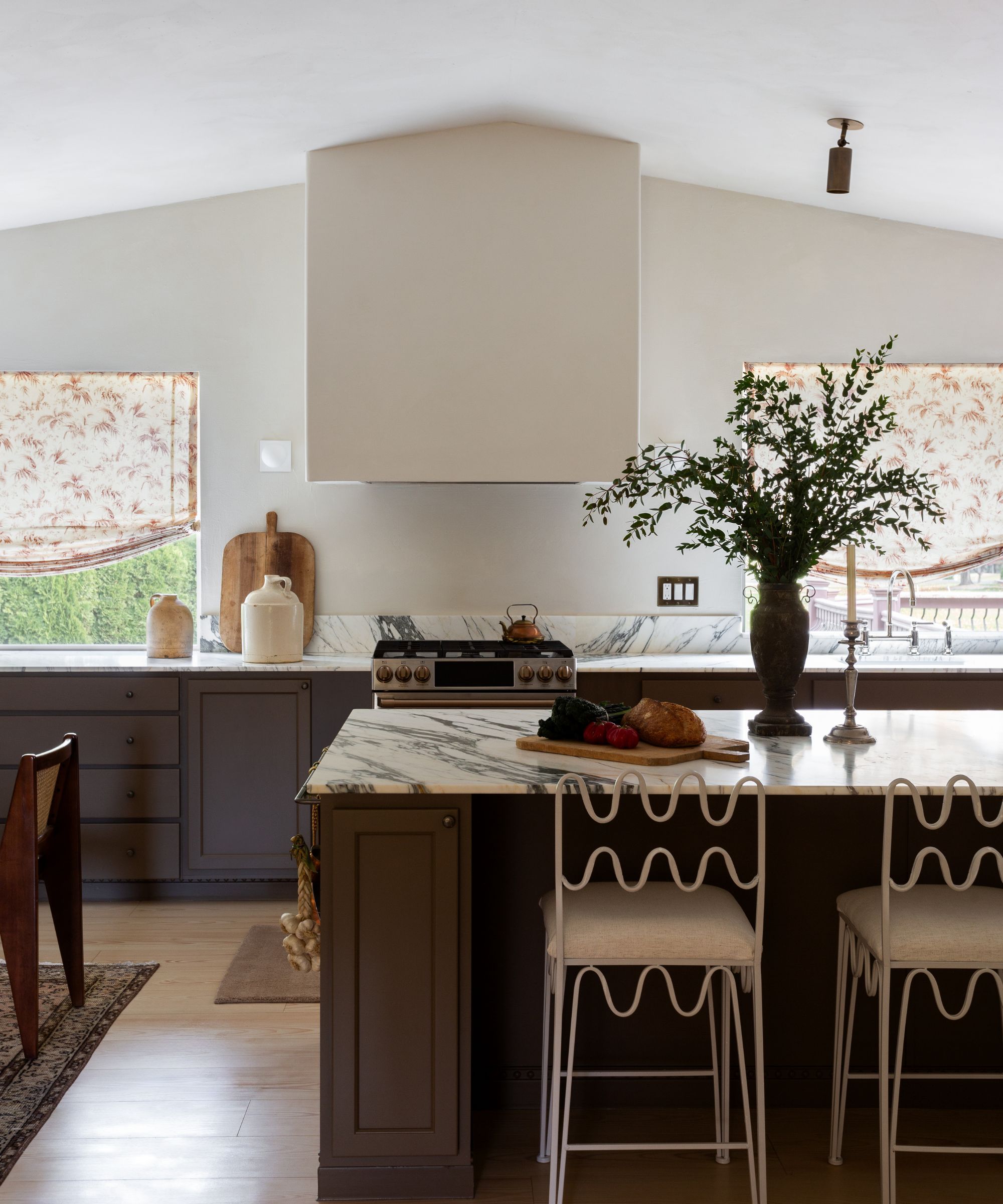 Kitchen with broccoli brown by Farrow & Ball painted island and cabinets, white marble countertops, wrought-iron wiggly white bar stools, a large oven hood, and floral cream blinds.