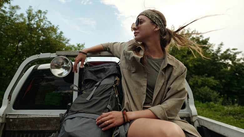 Young pretty female traveler riding in body of pickup truck and enjoying hitchhiking trip while looking at nature around