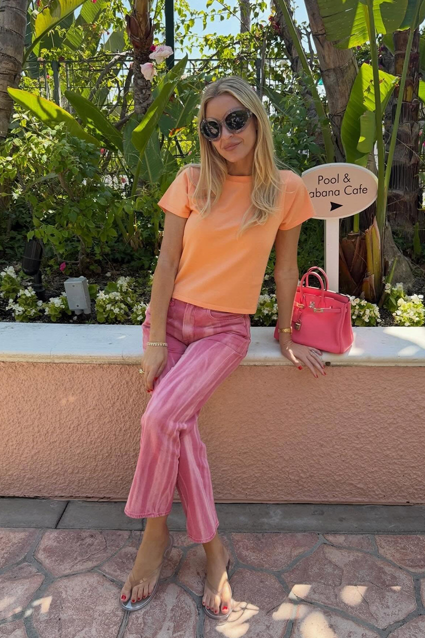 A woman wearing an orange T-shirt with pink striped pants, jelly sandals, oversize sunglasses, and a pink bag.