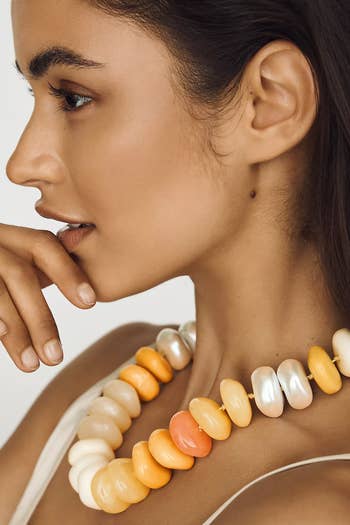 Model in profile wearing a chunky necklace with large, smooth beads of varying shades, contemplating with her hand near her face