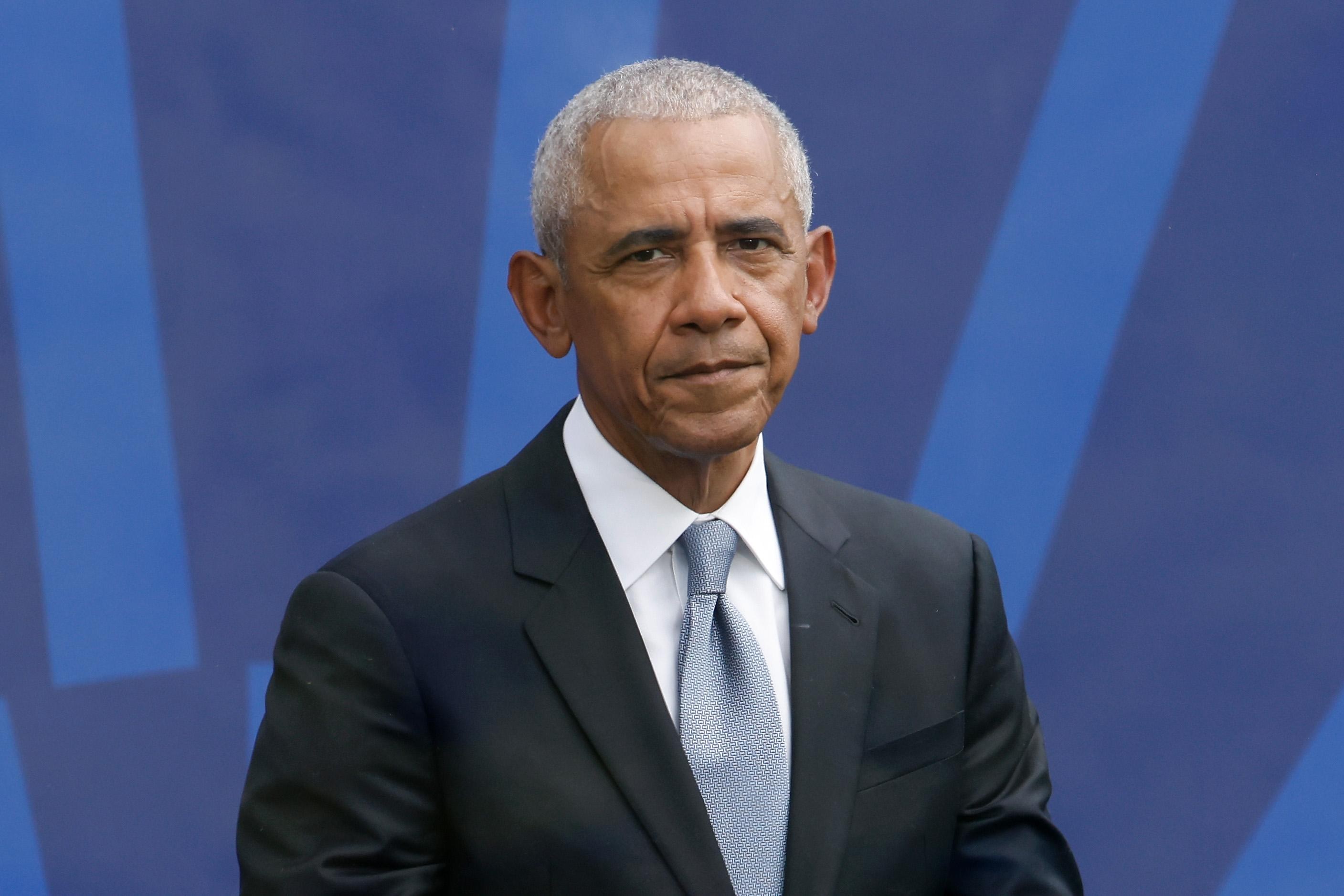 Former President Barack Obama wearing a black suit, light blue tie, and sitting against a blue background.