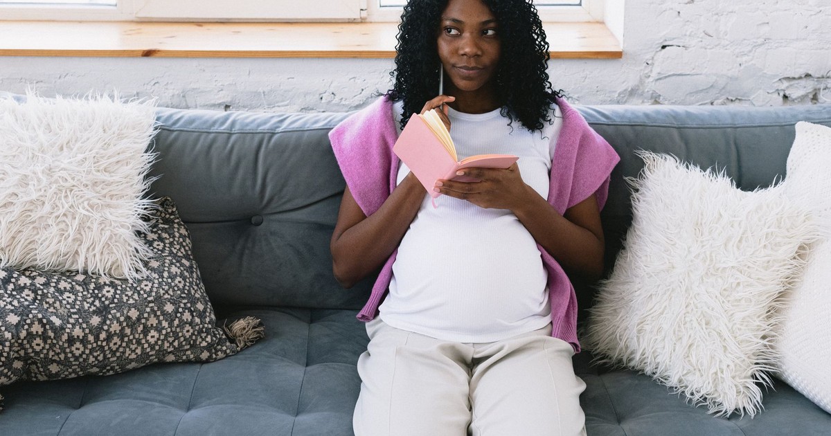 Thoughtful pregnant African American female taking notes in notepad and looking away while sitting on comfortable couch in living room