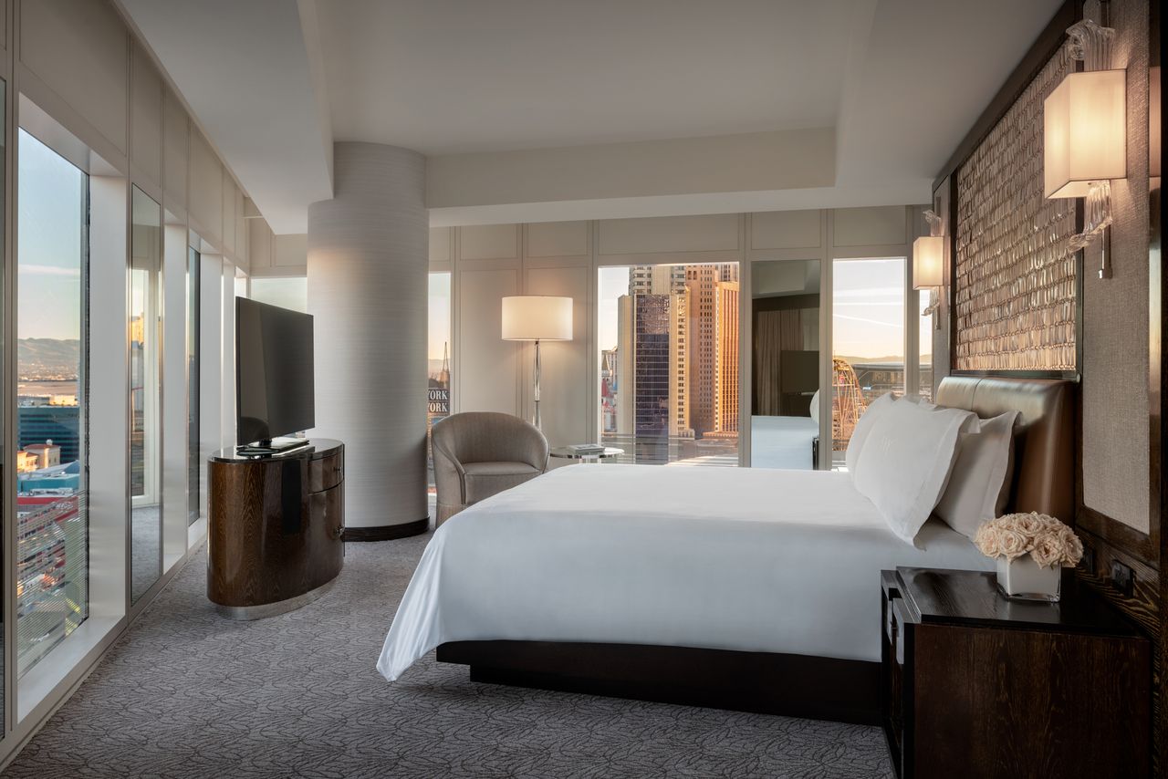Luxurious hotel room with a bed a corner chair TV and large windows offering a city view with buildings in the distance