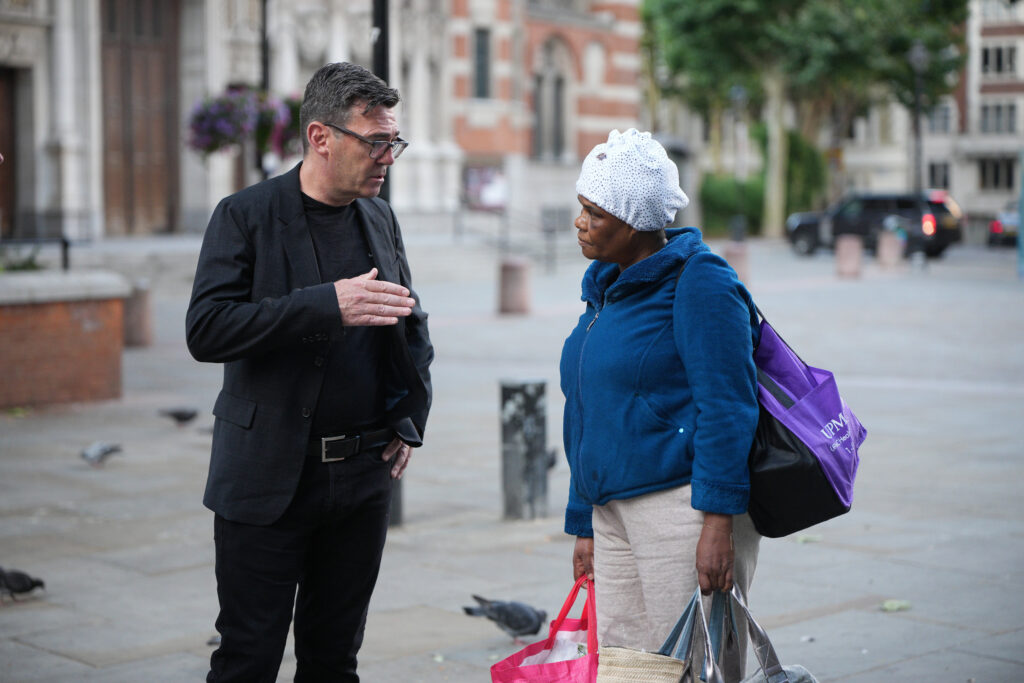 A man in a black blazer speaks with a woman carrying shopping bags on a city street