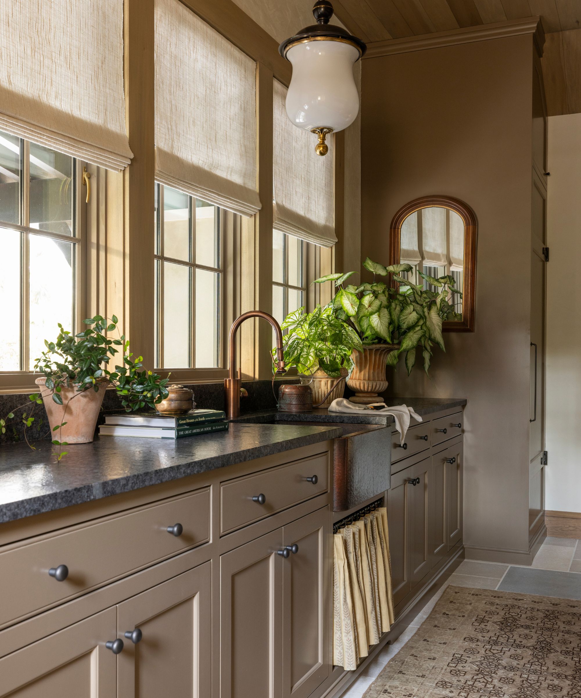 Mudroom with cabinetry and walls painted in Farrow & Ball's Broccoli brown, three large windows, a dark marble countertop, a sink with a skirted cupboard underneath, and plants in rustic pots.