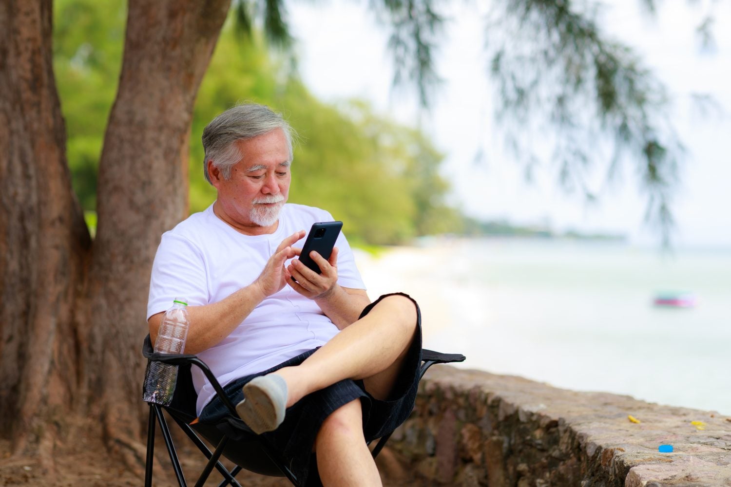 Asian senior male using mobile phone for video call communication with his family while vacation next to the beach.