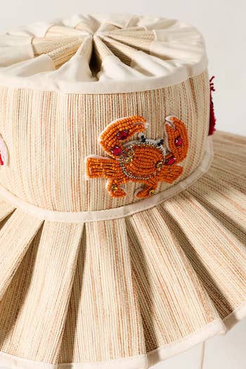 A pleated hat with an embroidered crab design, featuring decorative beads and gemstones, suitable for stylish outdoor wear