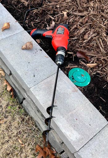 Garden auger attached to a drill, resting on stone ledge with mulch and bulbs nearby, suggesting use for planting