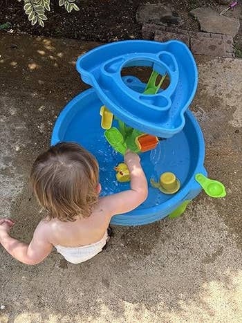 Child playing with a water play table outdoors, engaging with floating toys and cups, surrounded by grass and a concrete surface