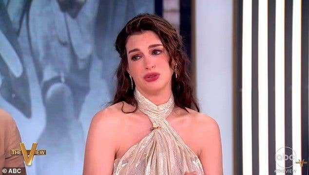 Anne Hathaway in a gold dress and scarf, speaking on a talk show set.