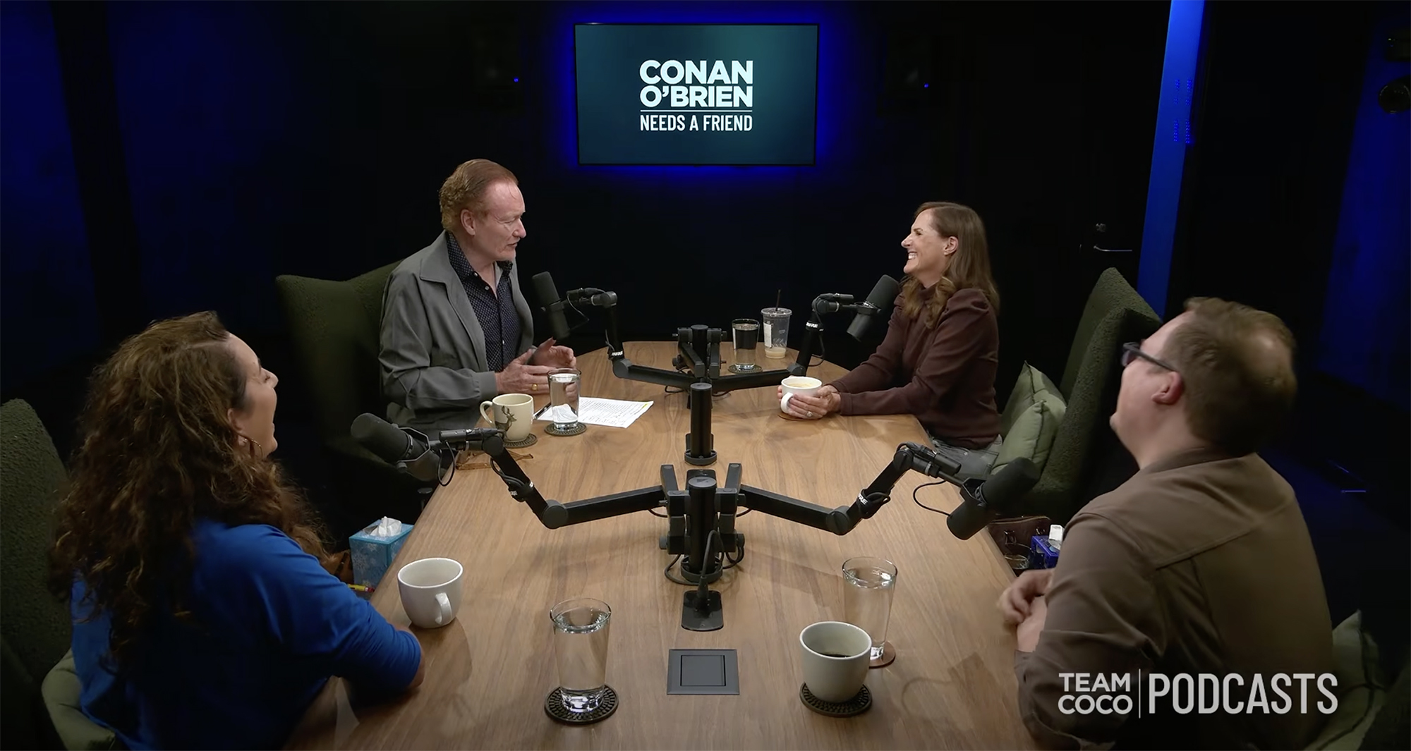 Conan O'Brien and three other people sitting around a table with microphones for a podcast.