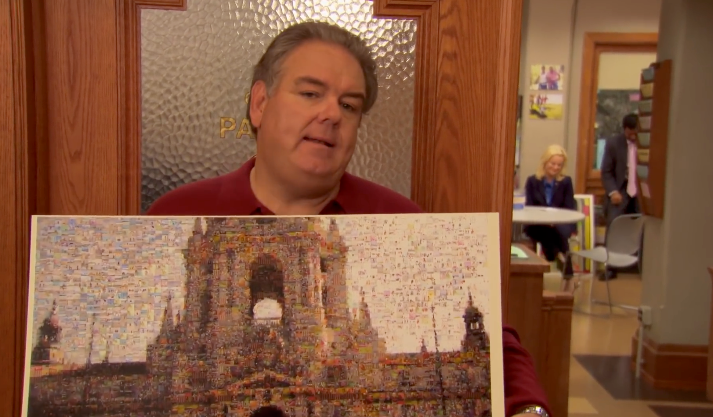 A person in an office holds a detailed mosaic portrait of a historic building. A woman and someone seated are visible in the background