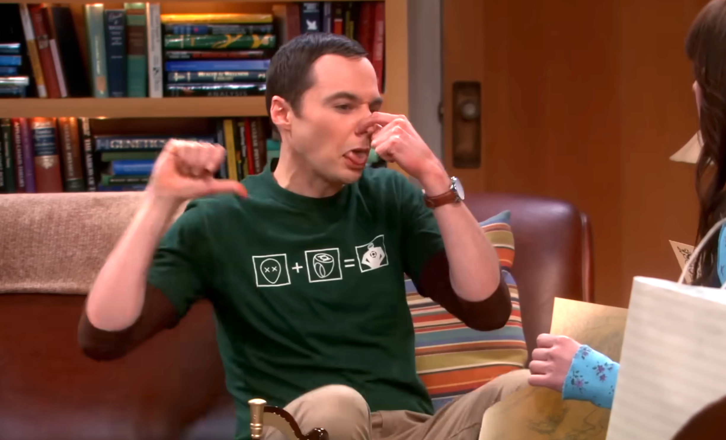A person playfully holds their nose while making a thumbs-down gesture, seated on a couch with bookshelves in the background