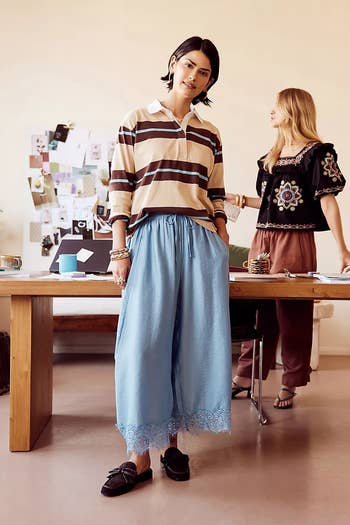 Two models in a relaxed office; one wears a striped shirt and blue wide-leg pants, the other in a boho blouse and rust pants, near a desk with papers
