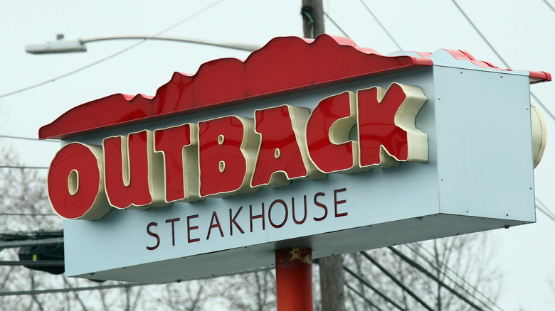 A light blue, red, and yellow Outback Steakhouse sign on a red pole during the day time.