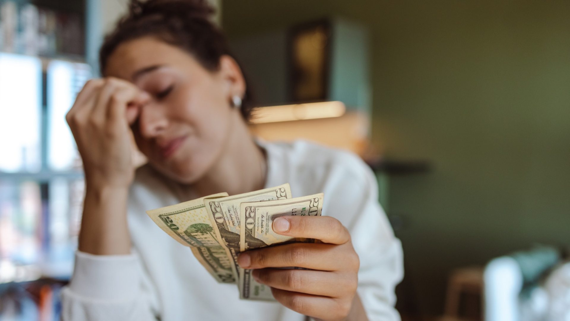 Discover a worried woman holding a few cash bills in her home while pinching the bridge of her nose