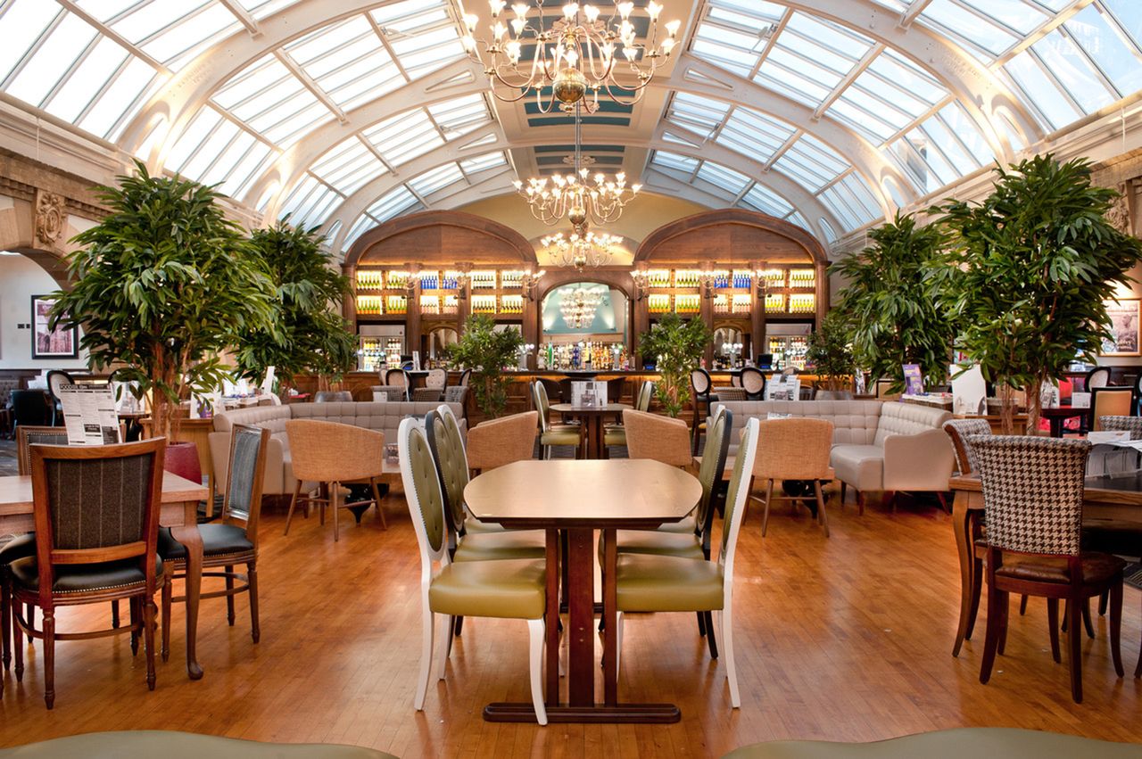 Interior of a grand dining and seating area showcasing wooden tables lush greenery and a large skylight ceiling