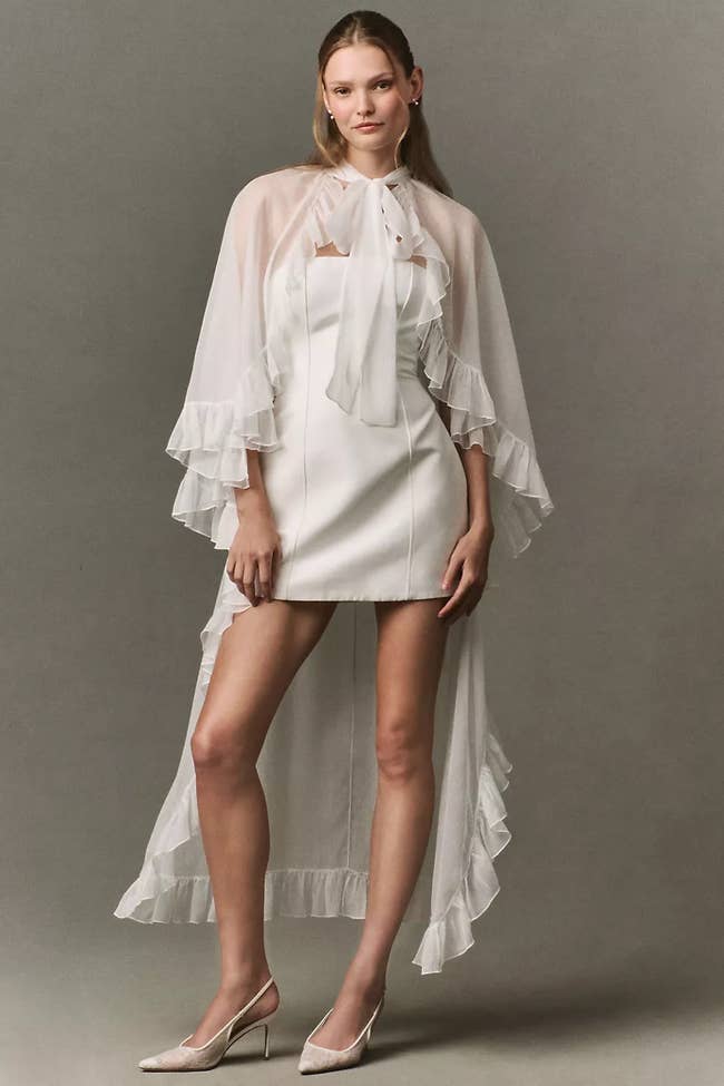 Model wearing a sheer cape dress with ruffled details over a short white dress, paired with heels