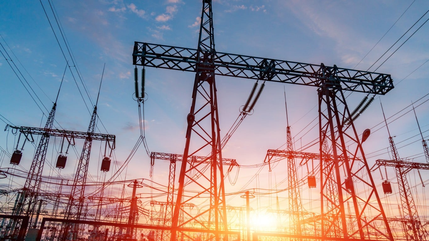 A low-angle shot of multiple electricity transmission towers and power lines silhouetted against a dramatic sky. The sky is deep blue at the top, transitioning to warm orange and yellow near the horizon, with a prominent sun glare in the lower right. The intricate steel lattice structures of the towers are clearly visible, with numerous insulators and high-voltage cables crisscrossing the frame.