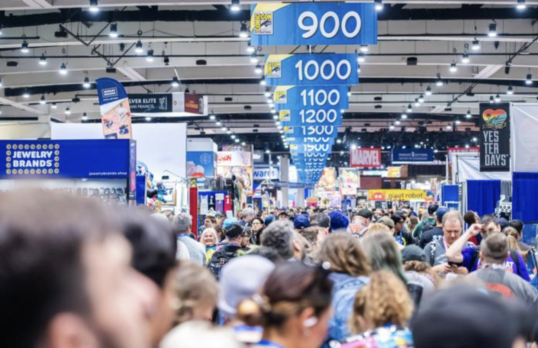 A crowd of people makes its way through a congested exhibit hall. Overhead are banners listing aisles by number, starting with