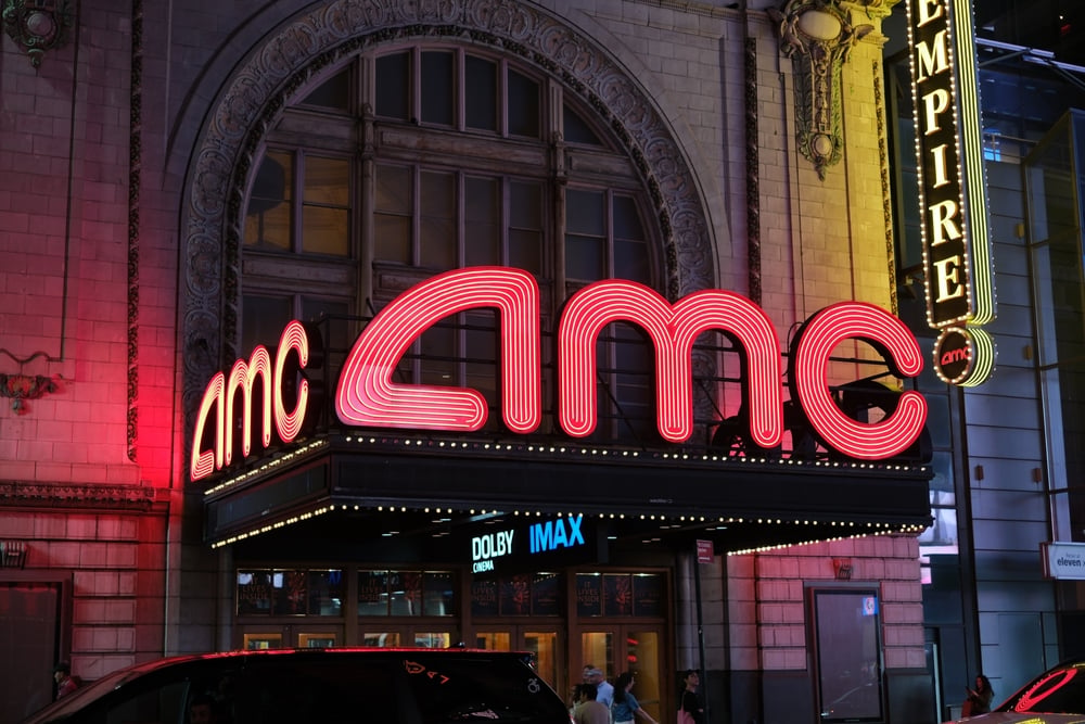 A large, red neon AMC sign illuminates an ornate theater entrance at night, with Dolby and IMAX logos.