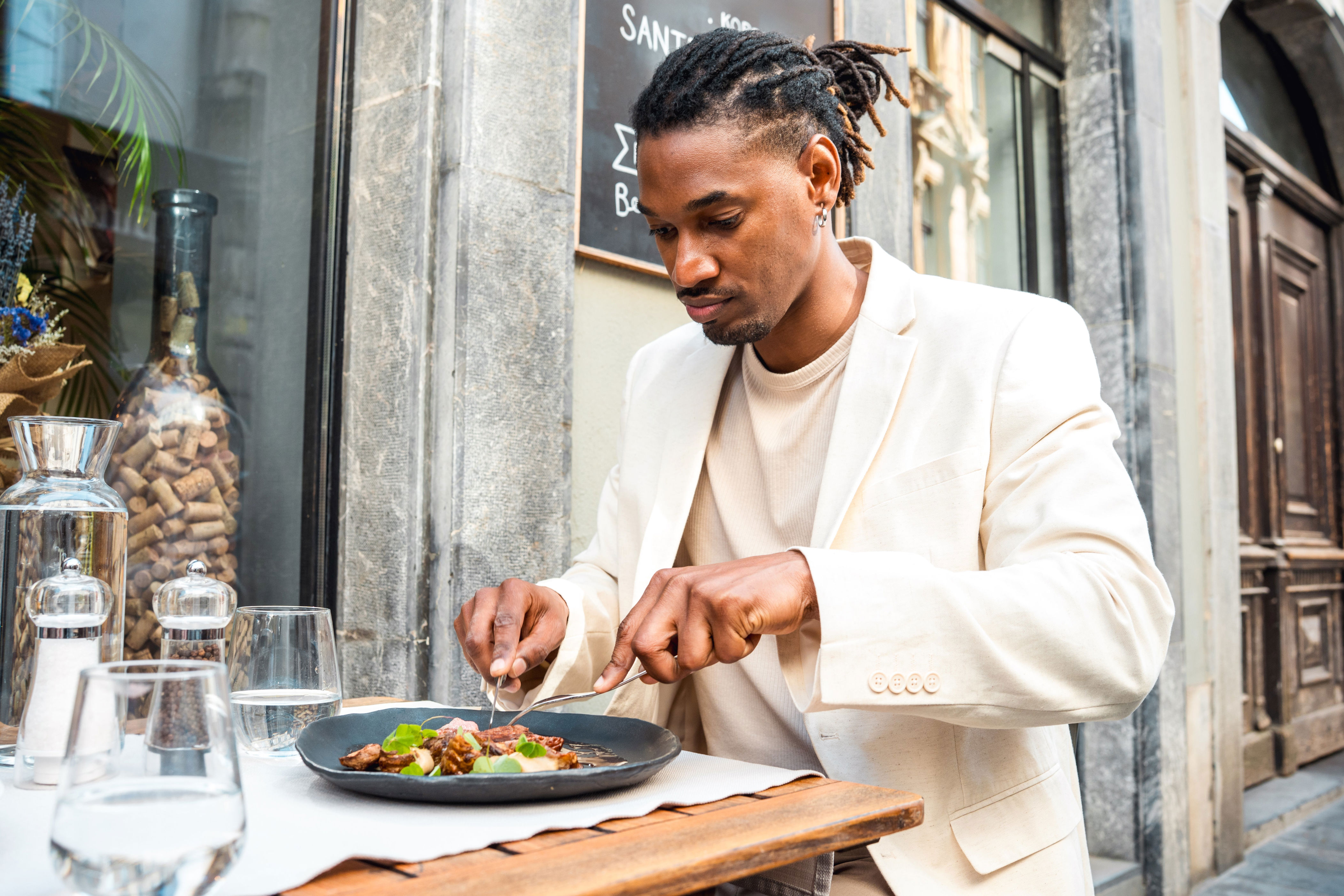Person in a light suit dining outdoors, using a fork and knife to enjoy a meal on a stylish plate at a restaurant terrace