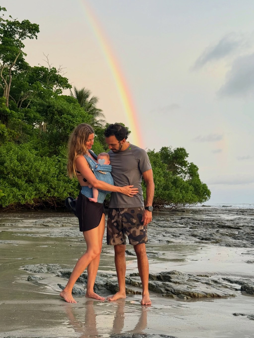 Gisele Bündchen and Joaquim Valente stand on a beach, holding a baby in a carrier, with a rainbow visible in the sky.