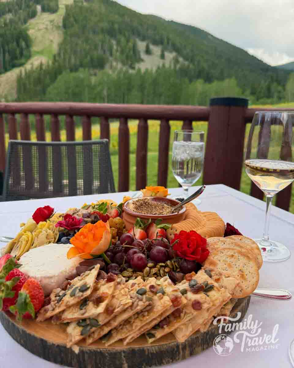 A charcuterie board with wine on a deck overlooking green mountain ski slopes, likely in a resort town.