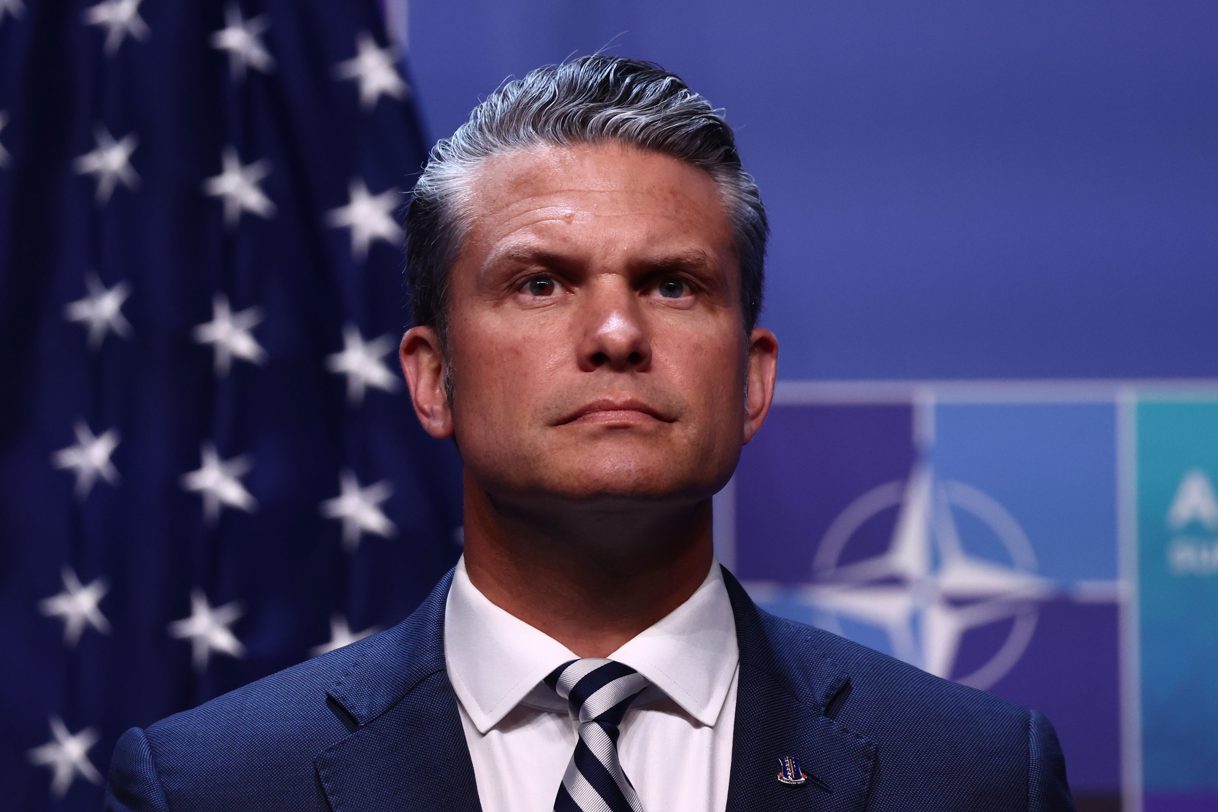 A man in a suit and tie stands in front of NATO flags, looking forward with a composed expression