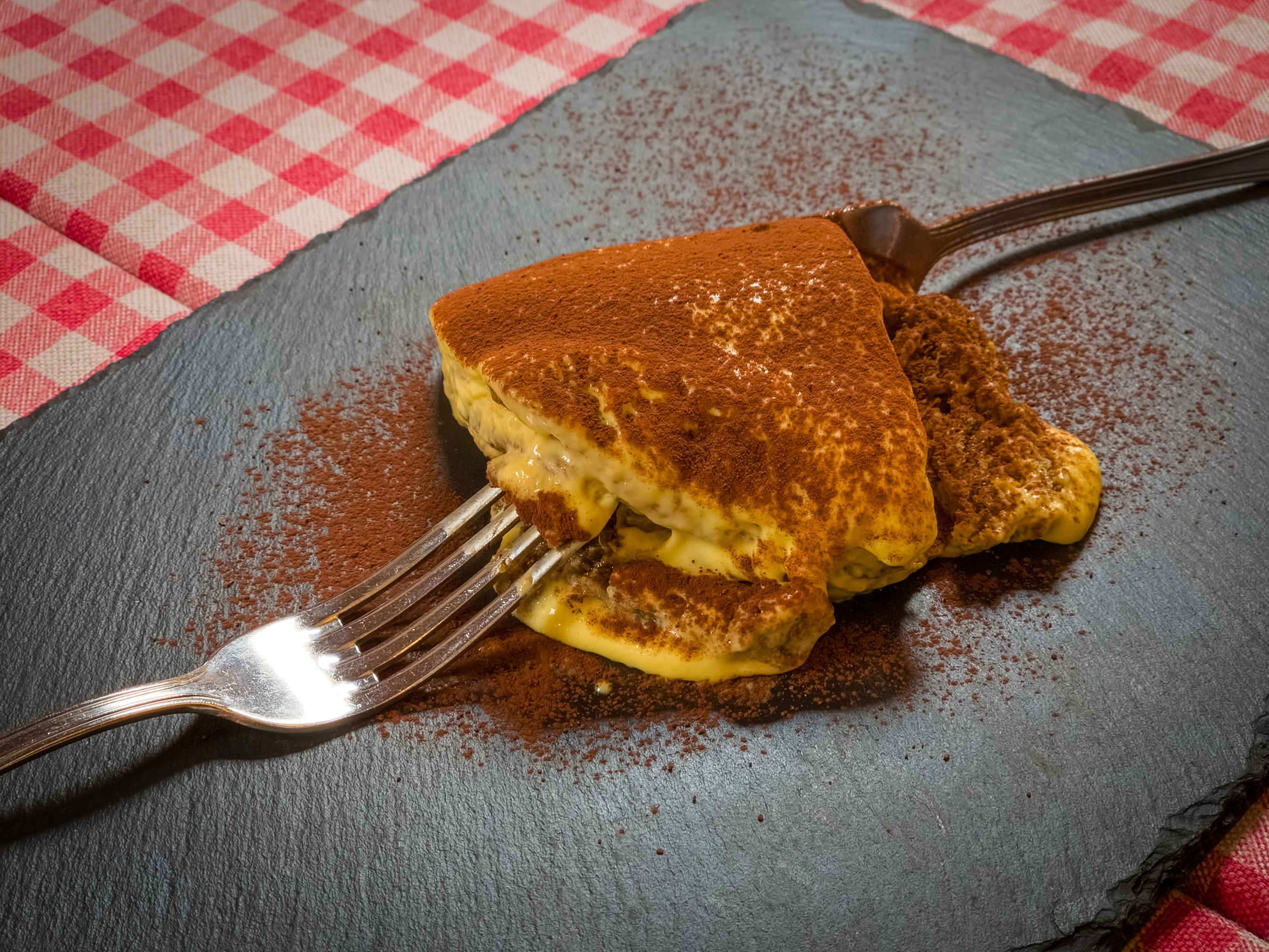 Slice of tiramisu on a slate plate with a fork on a checkered tablecloth