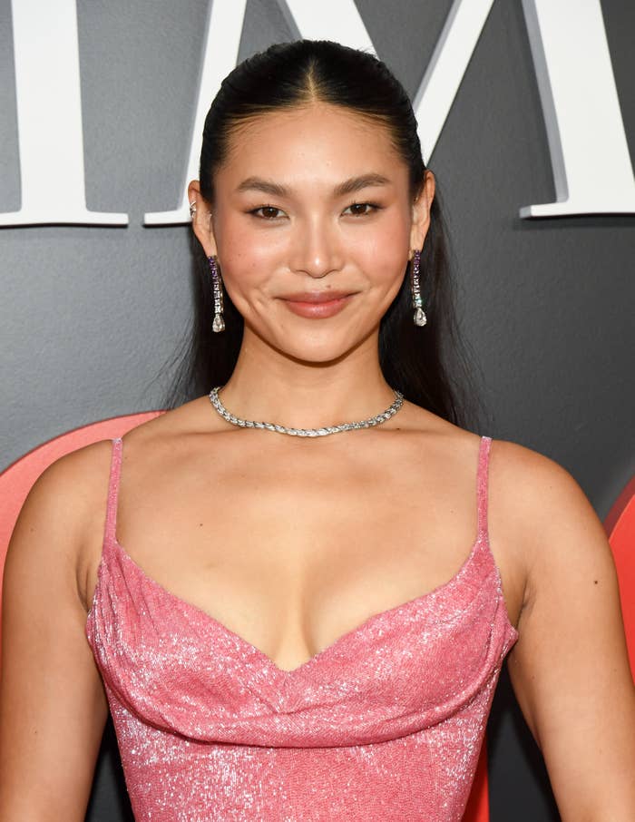 Person with long hair wearing a glittery gown and jewelry, posing on a red carpet event