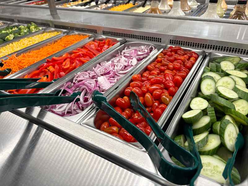 Fresh vegetables in a salad bar: red bell peppers, cucumbers, cherry tomatoes, shredded carrots, and red onions.